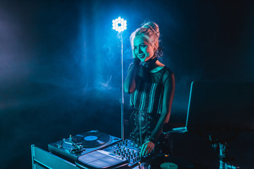 happy blonde dj girl in headphones standing near dj mixer in nightclub with smoke