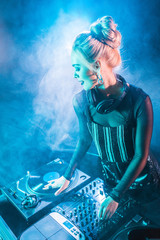 happy blonde dj girl in headphones standing near dj mixer and vinyl record in nightclub with smoke