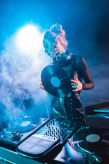 blonde dj woman looking at dj equipment and holding retro vinyl record in nightclub with smoke