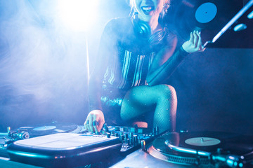 cropped view of cheerful dj woman holding retro vinyl record and standing near dj equipment in...