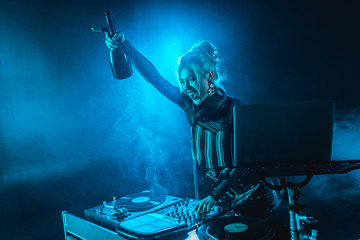 cheerful blonde dj woman in headphones holding  bottle and screaming near dj equipment in nightclub...