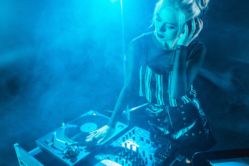 blonde dj girl listening music in headphones while looking at dj equipment in nightclub with smoke