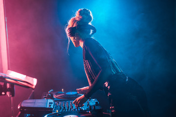 dj woman with blonde hair using dj mixer and touching vinyl record in nightclub with smoke