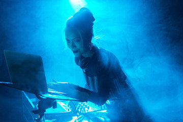 happy dj woman with blonde hair using laptop near dj equipment in nightclub with smoke