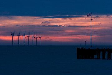 Colourful dawn sky with the sun rising over the sea behing an offshore wind farm.  Union jack flag flying at the end of a jetty