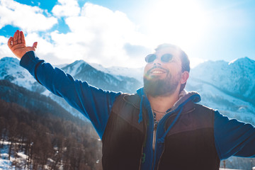 An adult man with a beard is standing on top of a mountain, smiling. Hands spread, a broad smile on his face. Enjoys a winter holiday in a ski resort