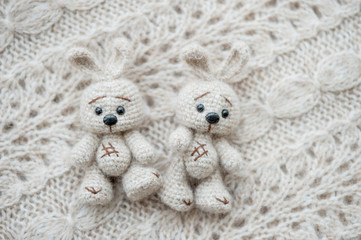 Two knitted white hares are lying on a light knitted handmade fabric. There is a free space.