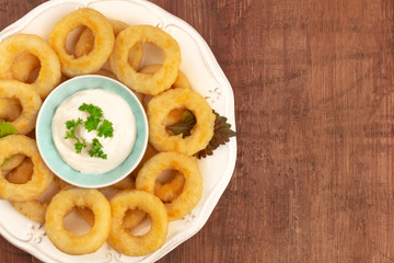 A closeup of a plate of squid rings with a mayonnaise dip, shot from the top on a dark rustic wooden background with a place for text