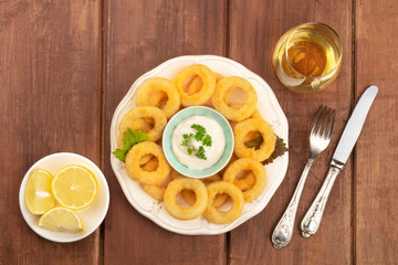 Squid rings with a mayonnaise dip, lemons, and a glass of wine, shot from the top on a dark rustic wooden background with copy space
