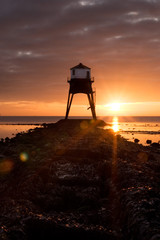 Old Victorian Gas Lightouse on shoreline with sunrise or sunset behind it.  Coastal landscape