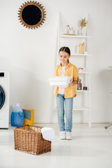 child in yellow shirt and jeans holding towels in front of ladder in laundry room