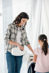 mother in grey shirt pouring liquid laundry detergent in washer wile daughter standing in laundry room