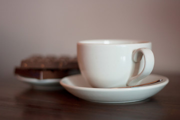 White cup on a saucer with metal spoon with chocolate biscuits in the background.  Coffee shop, tea shop, cuppa with friends, relaxing, take a break concepts.