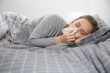 Sick woman lying in bed with closed eyes, blowing nose 