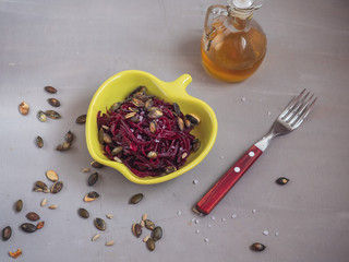 Grated boiled beets with sunflower and pumpkin seeds in a yellow ceramic salad bowl, in the background a bottle of vegetable oil.