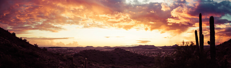 A panorama of a dramatic sunset over the desert with saguaro cactus and colorful clouds in the sky.