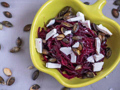 Salad of boiled beets and seeds, feta cheese in a yellow ceramic salad bowl, seasoned with vegetable oil. A wooden knife with a reddish wooden handle also rests on a wooden gray background. - Powered by Adobe