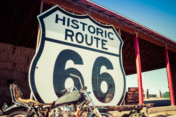 A large Route 66 road sign with a weathered motorcycle in the foreground. - obrazy, fototapety, plakaty