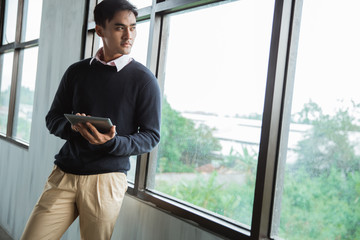 young asian businessman with a pad standing look out for the windows in the office