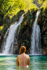 Young plump red-haired white woman with a bare back is standing in the turquoise water of a beautiful waterfall among the rocks in the jungle