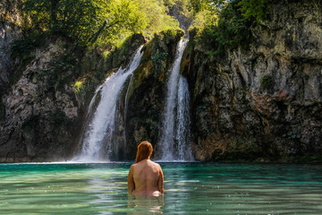 Fototapeta na wymiar Young plump red-haired white woman with a bare back is standing in the turquoise water of a beautiful waterfall among the rocks in the jungle
