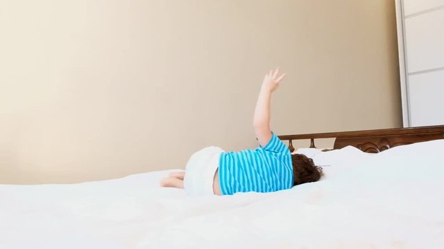 little boy in azure t-shirt plays on large comfortable parents bed jumping falling on back swinging legs slow motion