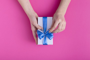 girl unpacks present in hands, women unties the blue ribbon bow on gift box on a colored pink cardboard background, top view, concept holiday, love and care
