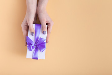 girl holding a present in hands, women with gift box with a tied lilac ribbon bow in hands on a pastel colored orange background, top view, concept holiday, love and care