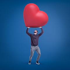 Young fit man in casual clothes holding up huge red Valentine heart on blue background.