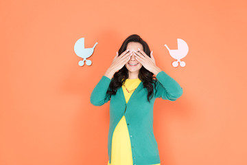 Glad pregnant woman in green cardigan covering eyes with hands on orange background