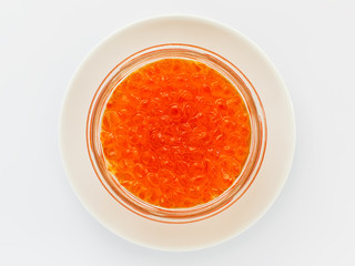 A glass jar of red caviar on a white porcelain plate on a white background. Salmon caviar. Sea fish delicacy.
