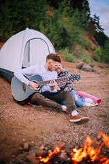 A man and a girl sit by the river and campfire and play the guitar.