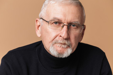 Old handsome man with gray hair and beard in eyeglasses and sweater thoughtfully looking in camera over beige background isolated