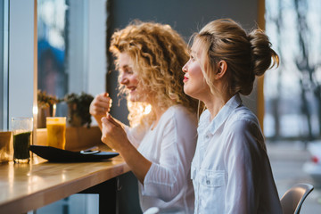 Two blond female friends talking and tasting healthy food drinking fresh smoothies at cafe or restaurant in evening time.