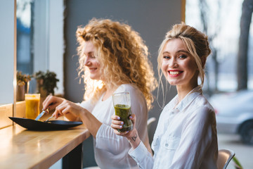 Two blond female friends talking and tasting healthy food drinking fresh smoothies at cafe or restaurant in evening time.