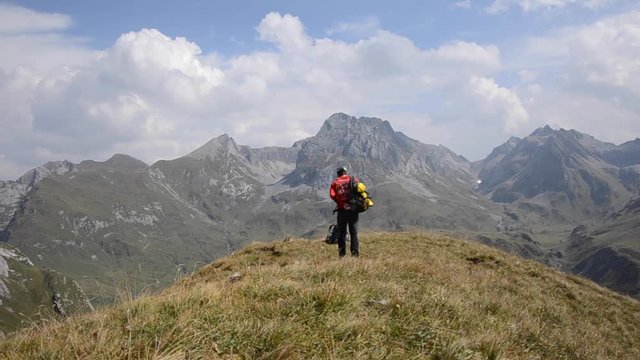 a male hiker walking away from camera in austrian alps in summer, then stopping and getting rid of his rucksack to take a break