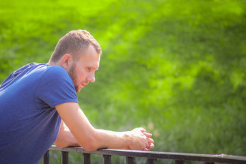 A young man in a blue T-shirt stands thoughtfully leaning on the railing in the park on a sunny summer day, blurry background