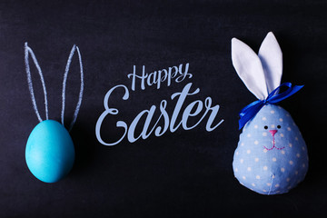 A blue painted Easter egg on a chalkboard with pressed ears looks like a rabbit. And the hare is handmade from fabric. Text, happy easter.