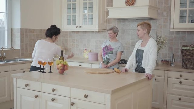 Three mature women communicating chatting at home standing near modern table in the middle of kitchen. Senior ladies having fun spending time together drinking wine and eating fruits