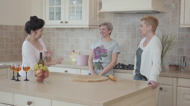 Adult girlfriends laugh and have fun at the table with fruit and wine in the kitchen. Elderly girlfriends have not seen each other for a long time and tell interesting stories to each other.