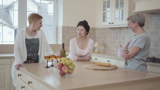 Elderly girlfriends have not seen each other for a long time and tell interesting stories. Adult girlfriends laugh and have fun at the table with fruit and wine in the kitchen.