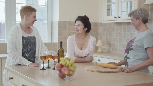 Elderly girlfriends have not seen each other for a long time and tell interesting stories. Adult girlfriends laugh and have fun at the table with fruit and wine in the kitchen.