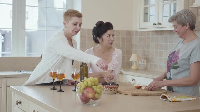Three middle aged mature women communicating chatting at home standing near modern table in the middle of kitchen. One lady cutting apple. Senior ladies having fun drinking wine and eating fruits