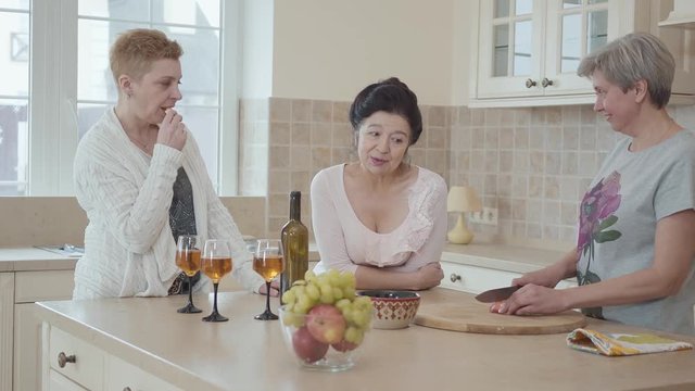 Mature women communicating chatting at home standing near modern table in the middle of kitchen. One lady cutting apple. Senior ladies having fun drinking wine and eating fruits
