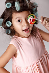 Funny little child girl in pink dress and hair curlers holding lollipop on gray background.