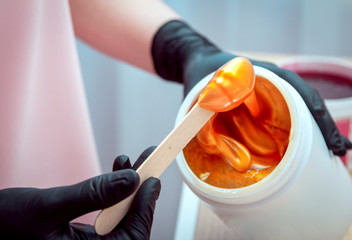 Close-up hands of cosmetologist in black gloves preparing golden colored waxing paste on spatula for sugaring depilation, hair removal beauty procedure.