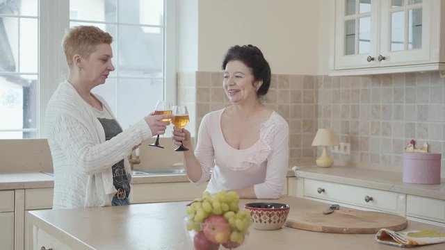 Mature women communicating at home standing near modern table in the kitchen. One lady says a toast with a genuine smile and they clink glasses. Senior ladies having fun drinking wine together