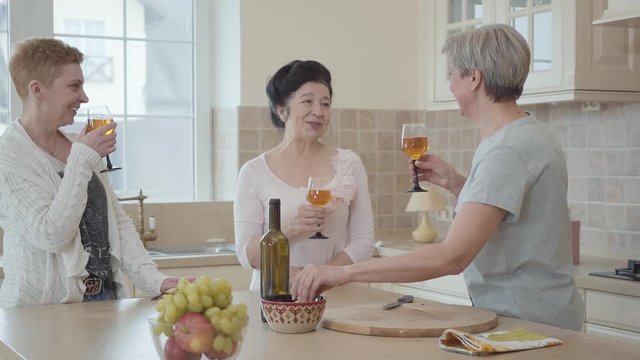 Three positive mature women communicating at home near modern table in the kitchen. One lady says a toast and they clink glasses. Senior ladies having fun drinking wine and eating fruits