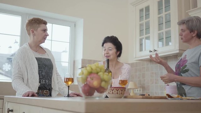 Adult cheerful girlfriends laugh and have fun chatting around the table with fruit and wine in the kitchen. Elderly girlfriends have not seen each other for a long time and are happy to talk.