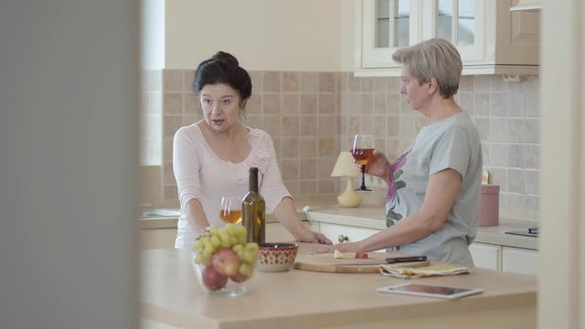 Two mature girlfriends met and chatting standing in the kitchen. Black-haired girl gesticulates and tells something to her friend. A gray-haired adult woman standing with a glass of wine.